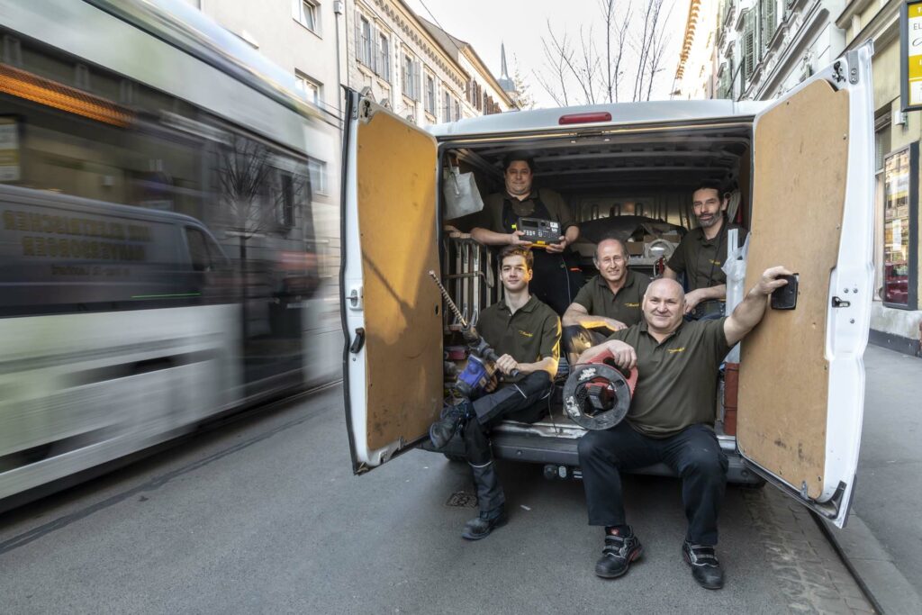 Mauerhofer-Team fuenf elektriker in montagewagen mit kabeltrommel schlagbohrer und stichsaege und vorbeifahrender strassenbahn