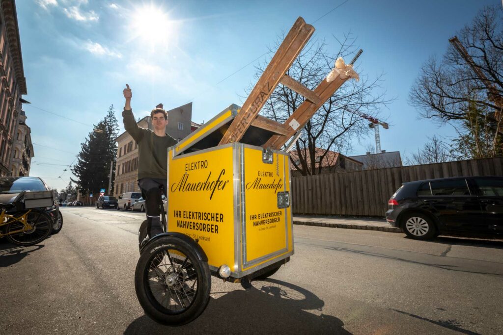 junger mitarbeiter von Elektro Mauerhofer auf dem gelben lastenrad mit firmenlogo transportiert eine leiter und winkt